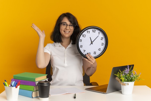 Smiling student with clock, books, and laptop on desk.